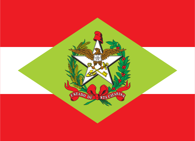 Três faixas horizontais, vermelha, branca e vermelha; no centro, um losango verde-claro com um brasão: uma águia sobre uma estrela branca, entre ramos.